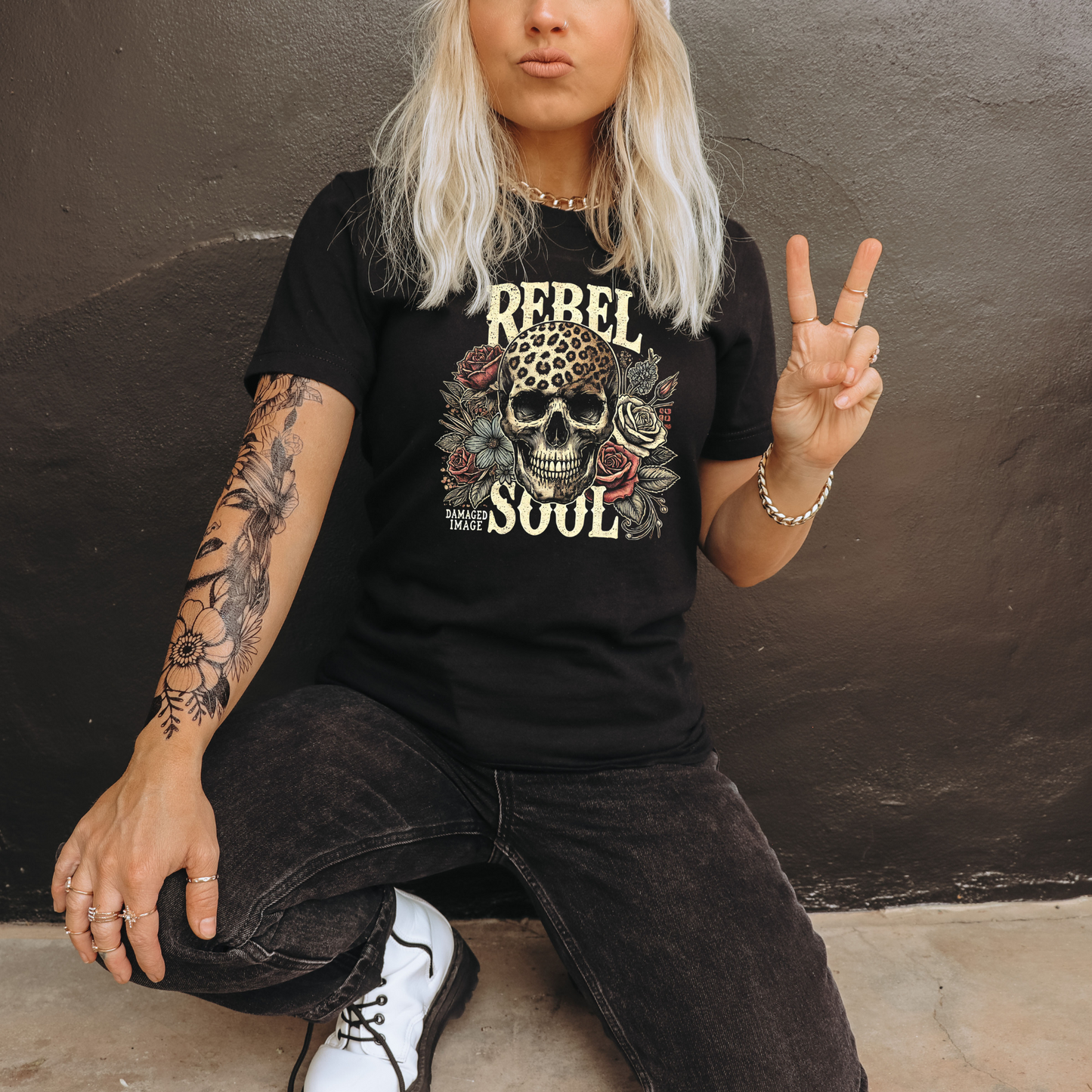 Person wearing a black t-shirt with a graphic design of a leopard skull and text that says rebel soul, sitting against a dark wall.
