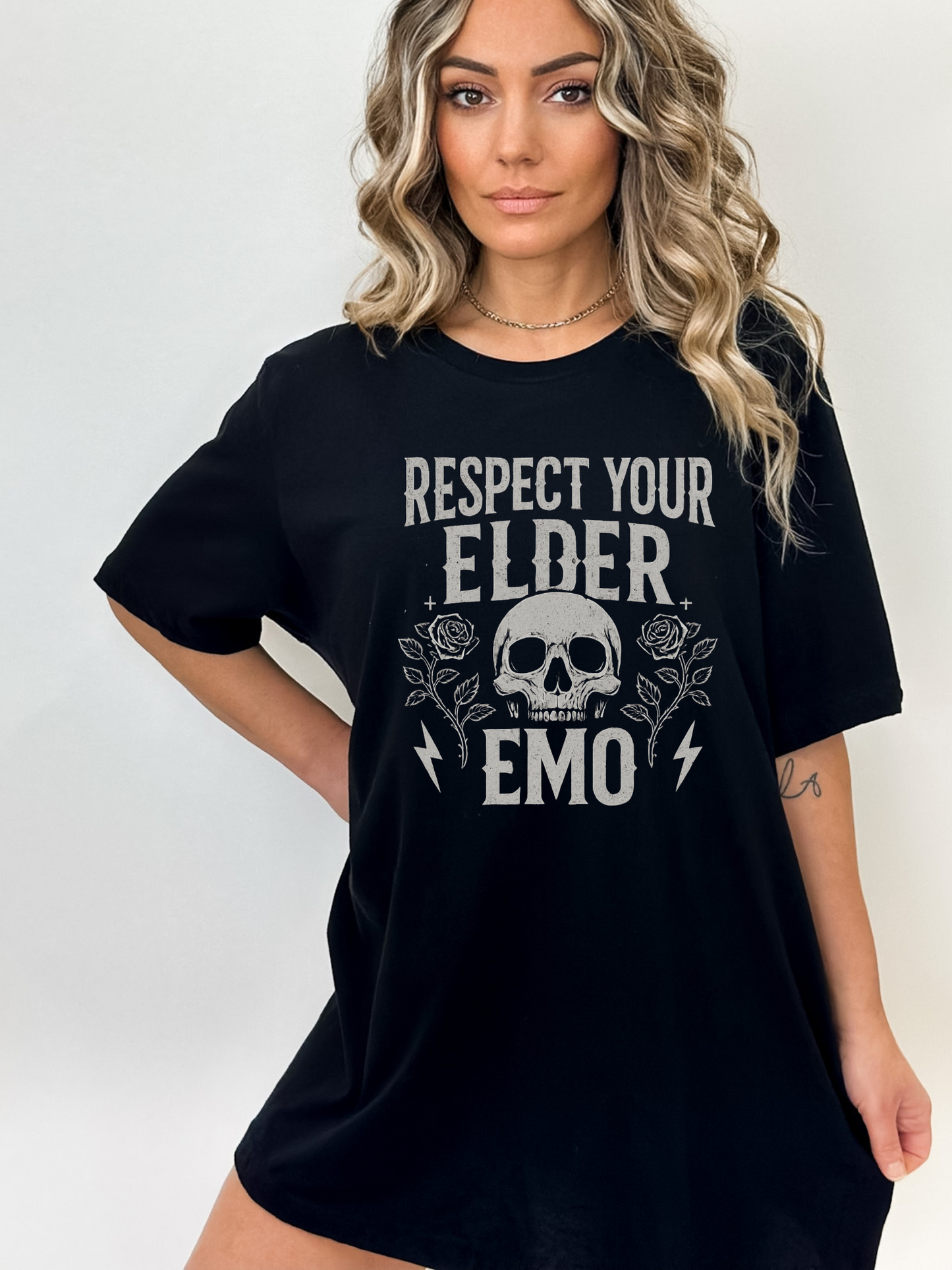 Woman wearing oversized black t-shirt with bold gothic text that reads "respect your elder emo" featuring a skull, roses, and lightning bolts. Styled with wavy blonde hair and a neutral background. 